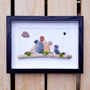 Op de afbeelding: Een ingelijst kiezelkunstwerk dat een gezin van vijf personen voorstelt die op een drijfhoutstam zitten. Het gezin is gemaakt van gladde, kleurrijke stenen, en een kleine hartvormige steen zweeft boven hen met een touwtje eraan vast. Het kunstwerk is geplaatst tegen een witte achtergrond.