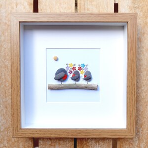 May include: A framed pebble art picture of three robins perched on a branch with colorful flowers behind them.
