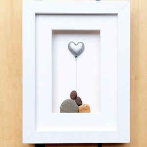 May include: A framed pebble art piece featuring a silver heart balloon floating above a stack of painted pebbles. The pebbles are brown, gray, and tan.