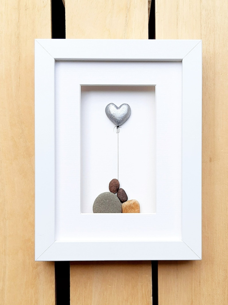 May include: A framed pebble art piece featuring a silver heart balloon floating above a stack of painted pebbles. The pebbles are brown, gray, and tan.