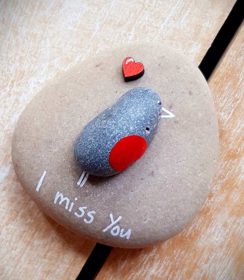 Puede incluir: Una piedra pintada de gris con un p&aacute;jaro pintado de rojo y las palabras "I miss you" escritas en pintura blanca. Un peque&ntilde;o coraz&oacute;n de madera rojo est&aacute; sobre el p&aacute;jaro.