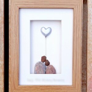 May include: A framed artwork featuring a silver heart-shaped balloon floating above three stacked stones. The stones are brown and gray, and the background is white.