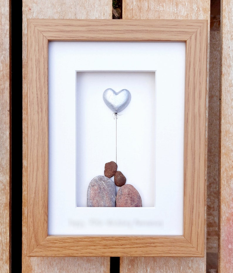 May include: A framed artwork featuring a silver heart-shaped balloon floating above three stacked stones. The stones are brown and gray, and the background is white.