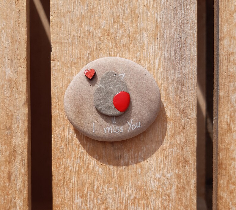 Puede incluir: Una piedra gris pintada con un petirrojo y las palabras "Te echo de menos". Un peque&ntilde;o coraz&oacute;n rojo est&aacute; pintado sobre el petirrojo.