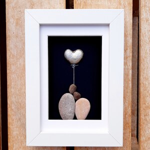 May include: A framed pebble art piece depicting a couple holding a silver heart balloon. The artwork is set against a black background and framed in white.