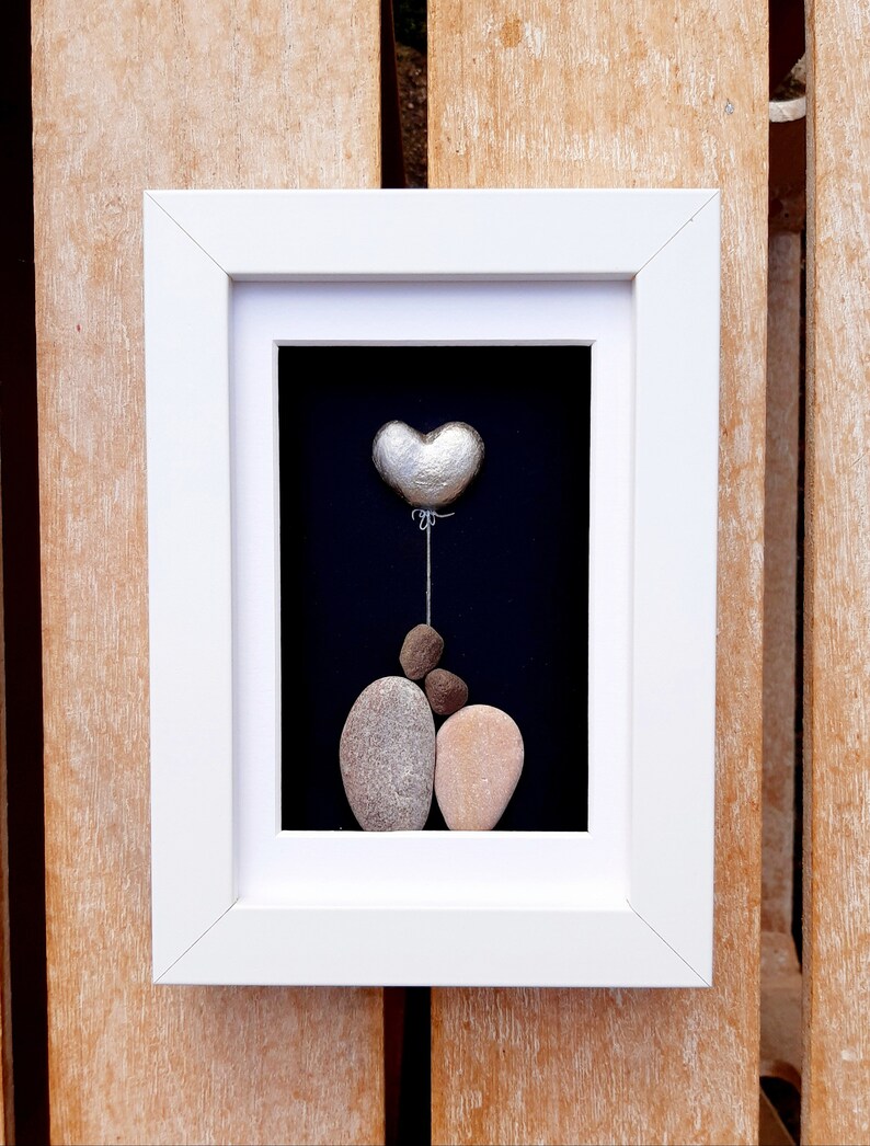 May include: A framed pebble art piece depicting a couple holding a silver heart balloon. The artwork is set against a black background and framed in white.