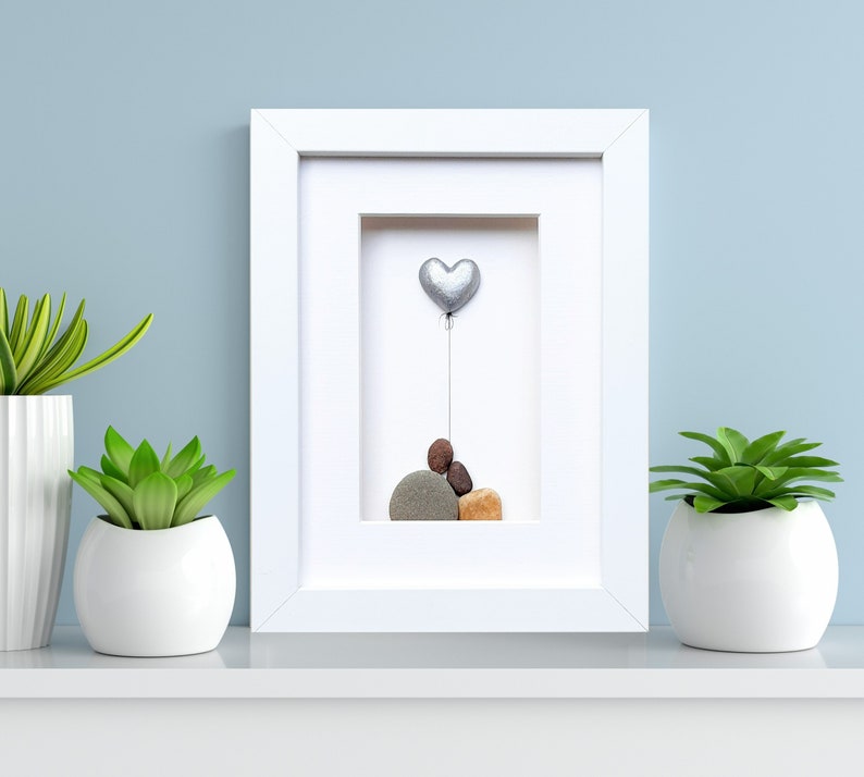 May include: A white frame with a white mat surrounding a pebble art piece. The art piece features a silver heart balloon floating above a stack of gray, brown, and tan pebbles.