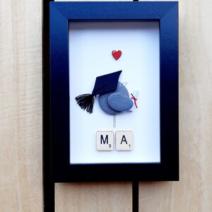 May include: A framed pebble art piece depicting a bird wearing a graduation cap and holding a diploma. The bird is made of gray pebbles and the cap is black. The background is white with a red heart at the top. The letters "M A" are spelled out with Scrabble tiles below the bird.