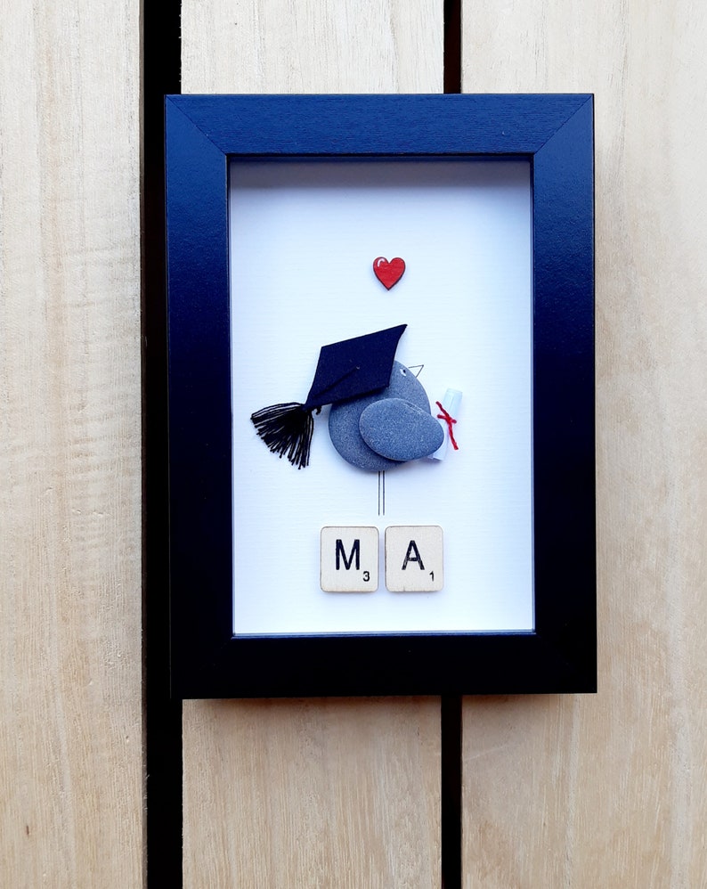 May include: A framed pebble art piece depicting a bird wearing a graduation cap and holding a diploma. The bird is made of gray pebbles and the cap is black. The background is white with a red heart at the top. The letters "M A" are spelled out with Scrabble tiles below the bird.