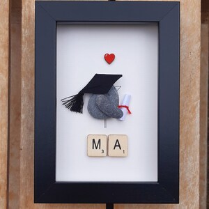 May include: A framed artwork featuring a gray stone painted to look like a graduation cap and gown, holding a scroll. The stone is topped with a black graduation cap. The artwork also includes a red heart and the letters "M.A." spelled out with Scrabble tiles.