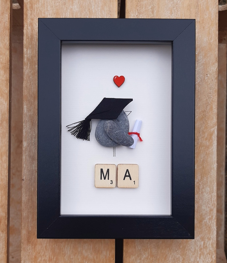May include: A framed artwork featuring a gray stone painted to look like a graduation cap and gown, holding a scroll. The stone is topped with a black graduation cap. The artwork also includes a red heart and the letters "M.A." spelled out with Scrabble tiles.