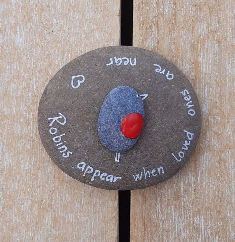 May include: A gray stone with white lettering that reads "When loved ones are near, Robins appear". A small painted bird with a red breast is in the center of the stone.