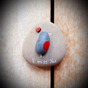 Puede incluir: Una piedra pintada de gris con un coraz&oacute;n rojo y un p&aacute;jaro pintado en ella. La piedra tiene las palabras "Te echo de menos" escritas en ella.