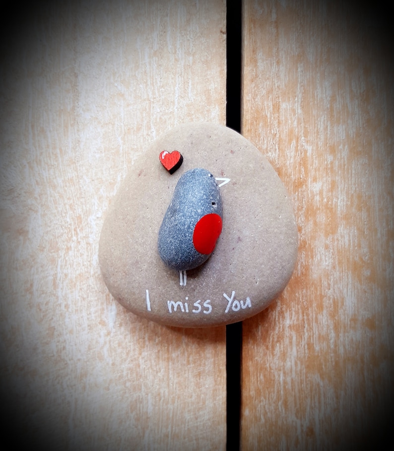 Puede incluir: Una piedra pintada de gris con un coraz&oacute;n rojo y un p&aacute;jaro pintado en ella. La piedra tiene las palabras "Te echo de menos" escritas en ella.