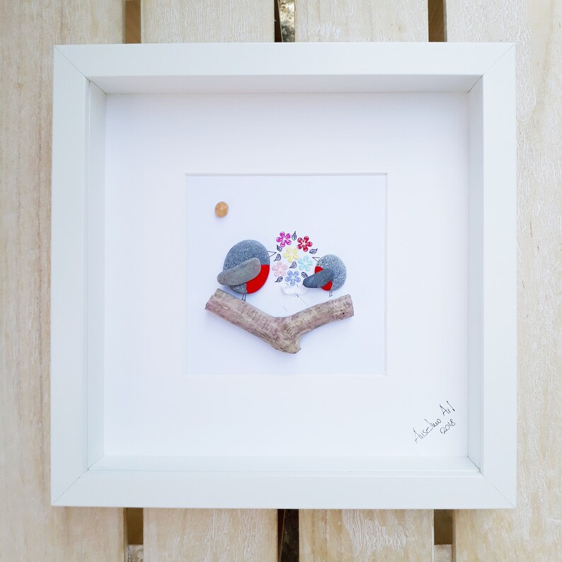 May include: Framed pebble art featuring two gray birds with red breasts perched on a branch. The birds are holding a bouquet of colorful flowers. The artwork is signed "Aliceda Art" and dated "2018".