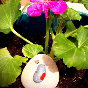 Puede incluir: Una piedra pintada con un dise&ntilde;o de p&aacute;jaro y las palabras "Te echo de menos". La piedra est&aacute; rodeada de hojas verdes y flores rosas.