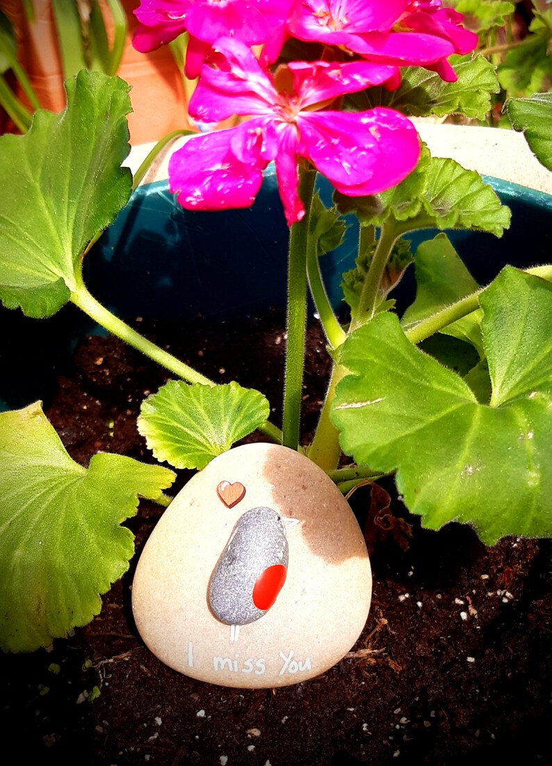 Puede incluir: Una piedra pintada con un dise&ntilde;o de p&aacute;jaro y las palabras "Te echo de menos". La piedra est&aacute; rodeada de hojas verdes y flores rosas.