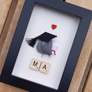 May include: A framed graduation pebble art piece with a black graduation cap, a red heart, and the letters "MA" spelled out with Scrabble tiles.