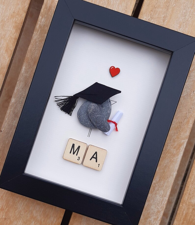May include: A framed graduation pebble art piece with a black graduation cap, a red heart, and the letters "MA" spelled out with Scrabble tiles.