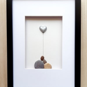 May include: A framed pebble art piece with a silver heart balloon floating above a stack of gray, brown, and beige pebbles.