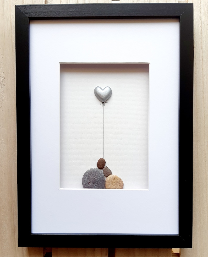 May include: A framed pebble art piece with a silver heart balloon floating above a stack of gray, brown, and beige pebbles.