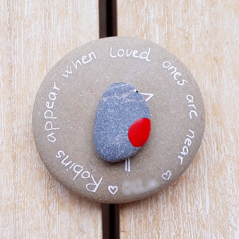 May include: A gray painted stone with the text "Robins appear when loved ones are near" and a painted robin with a red breast.