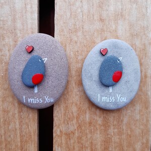Puede incluir: Dos piedras grises pintadas con corazones rojos y un p&aacute;jaro de pecho rojo. Las piedras tienen las palabras "Te echo de menos" escritas en ellas.