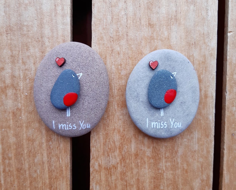 Puede incluir: Dos piedras grises pintadas con corazones rojos y un p&aacute;jaro de pecho rojo. Las piedras tienen las palabras "Te echo de menos" escritas en ellas.
