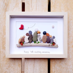 May include: A framed pebble art piece depicting a family of 10 people sitting on a driftwood log. A red heart is flying in the air above them. The text "Happy 40th wedding anniversary" is written below the family.