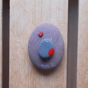 Puede incluir: Una piedra pintada de gris con un coraz&oacute;n rojo y un p&aacute;jaro gris con una mancha roja en el pecho. La piedra tiene las palabras "Te echo de menos" escritas en ella.
