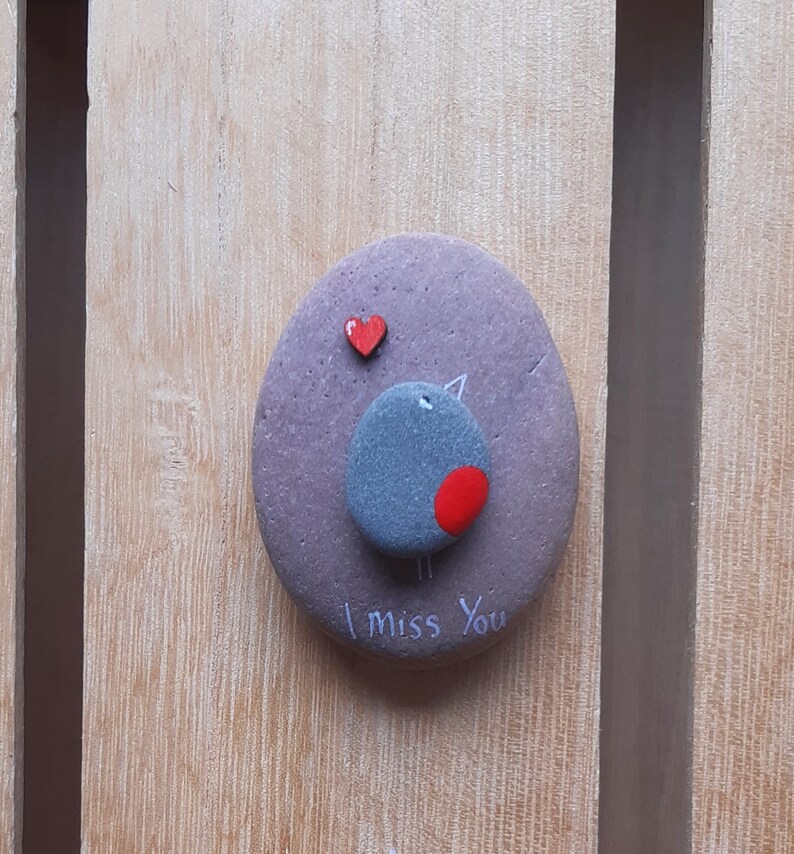 Puede incluir: Una piedra pintada de gris con un coraz&oacute;n rojo y un p&aacute;jaro gris con una mancha roja en el pecho. La piedra tiene las palabras "Te echo de menos" escritas en ella.