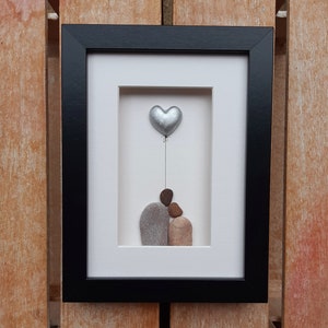 May include: A framed pebble art piece depicting a couple holding a silver heart balloon. The couple is made of gray and brown pebbles, and the heart is silver.
