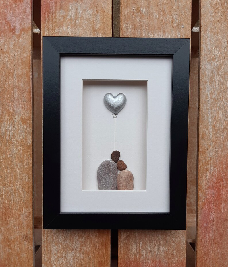 May include: A framed pebble art piece depicting a couple holding a silver heart balloon. The couple is made of gray and brown pebbles, and the heart is silver.