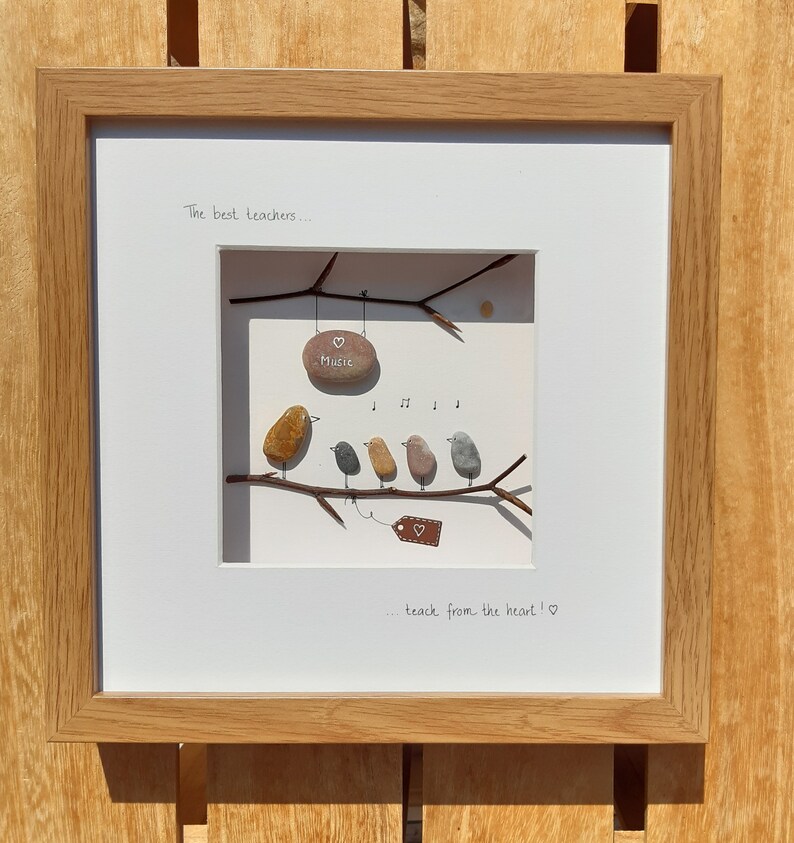 May include: Framed artwork featuring a hand-painted pebble art scene of five birds perched on a branch. The birds are singing and a sign that says "Music" hangs from the branch. The text "The best teachers... teach from the heart!" is written below the scene.