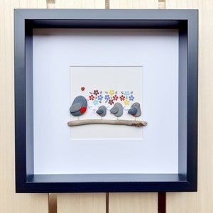 May include: A framed pebble art piece depicting a family of robins perched on a branch. The robins are made of gray and white pebbles, with the parent robin having a red breast. The background features colorful flowers and leaves.
