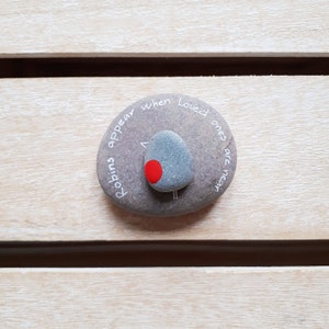 May include: A gray stone with the words "Robins appear when loved ones are near" written on it. A small gray stone painted to look like a robin with a red breast is on top of the larger stone.
