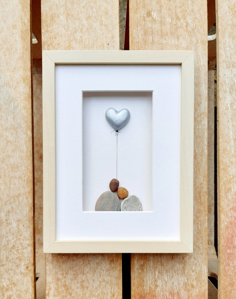 May include: A framed artwork featuring a silver heart-shaped balloon floating above a stack of gray, brown, and orange stones.