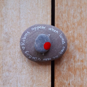 May include: A gray stone with the words "Robins appear when loved ones are near" written on it. A small gray stone with a red dot is painted on the stone to look like a robin.
