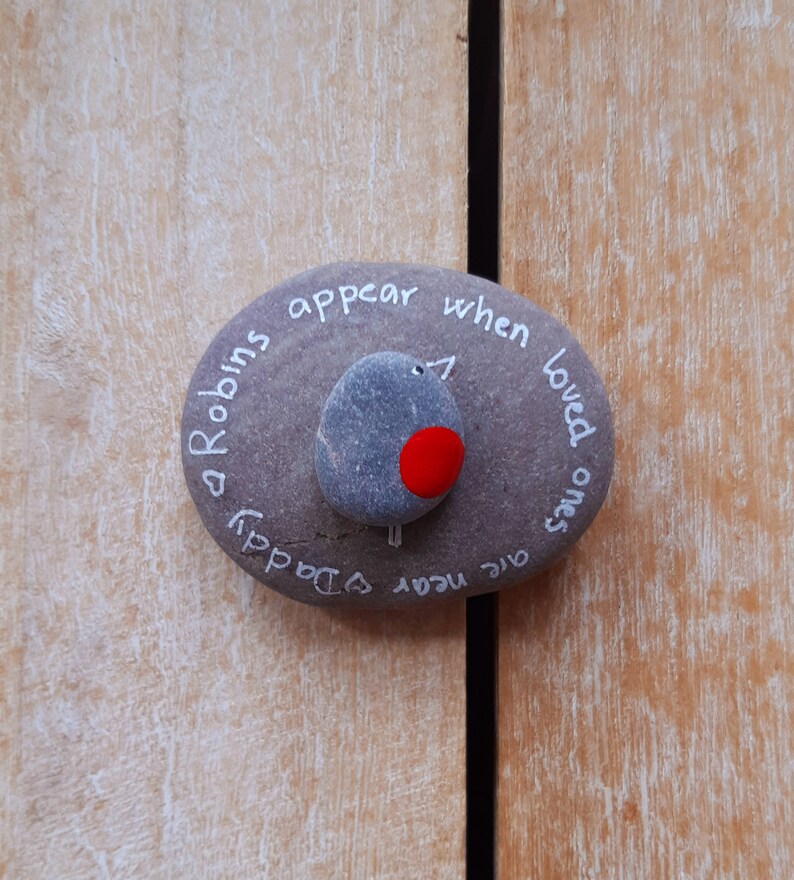 May include: A gray stone with the words "Robins appear when loved ones are near" written on it. A small gray stone with a red dot is painted on the stone to look like a robin.