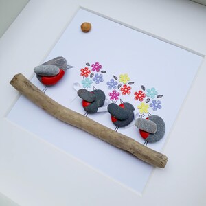 May include: Four gray stone robins with red bellies are perched on a branch. They are holding colorful paper flowers in their beaks. The robins are arranged in a line, facing the right side of the image. The background is white.