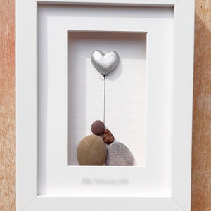 May include: A white framed picture with a silver heart shaped balloon and a pile of brown, gray, and red stones inside.