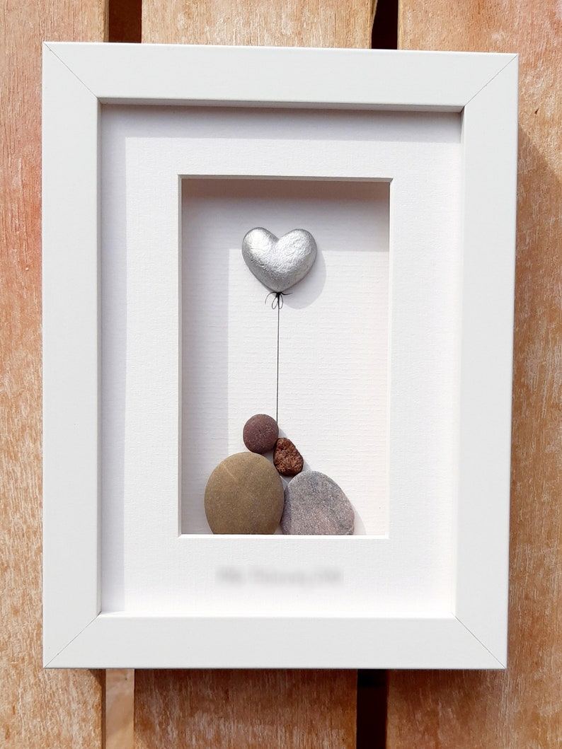 May include: A white framed picture with a silver heart shaped balloon and a pile of brown, gray, and red stones inside.