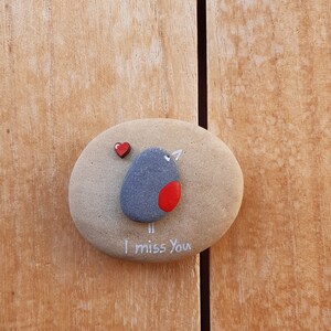 Puede incluir: Una piedra pintada de gris y rojo con un dise&ntilde;o de p&aacute;jaro y las palabras "Te echo de menos". Un peque&ntilde;o coraz&oacute;n rojo est&aacute; pintado en la piedra.