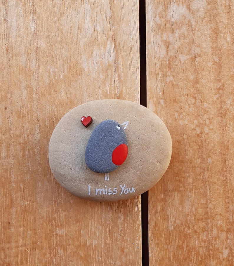Puede incluir: Una piedra pintada de gris y rojo con un dise&ntilde;o de p&aacute;jaro y las palabras "Te echo de menos". Un peque&ntilde;o coraz&oacute;n rojo est&aacute; pintado en la piedra.