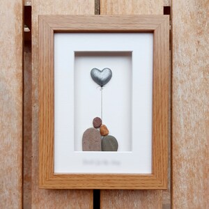 May include: A framed pebble art picture with a silver heart balloon and a stack of pebbles in the shape of a person. The frame is made of wood and has a white mat.