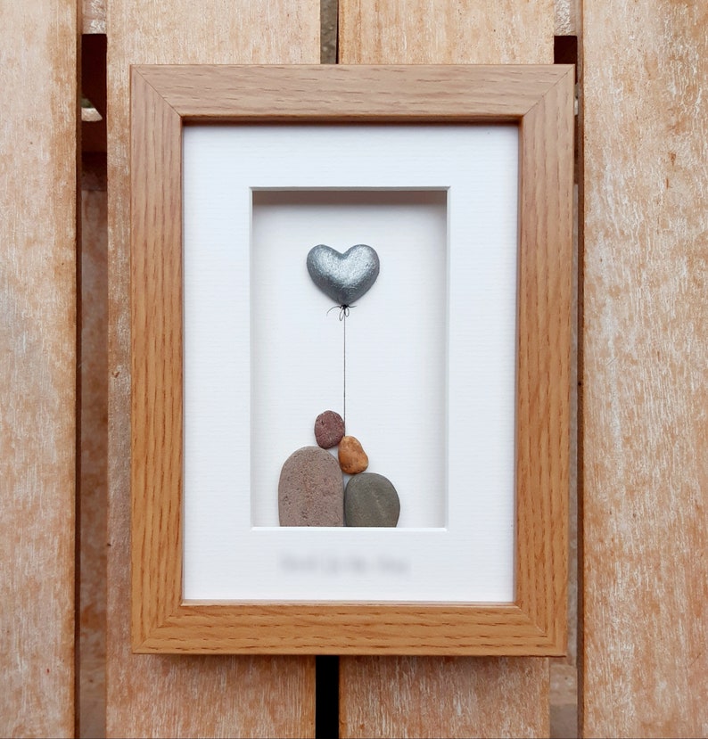 May include: A framed pebble art picture with a silver heart balloon and a stack of pebbles in the shape of a person. The frame is made of wood and has a white mat.