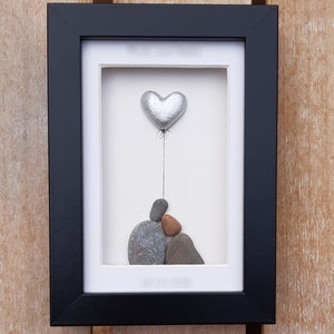 May include: A framed pebble art piece with a silver heart balloon floating above a stack of gray and brown pebbles.