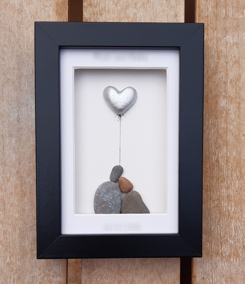 May include: A framed pebble art piece with a silver heart balloon floating above a stack of gray and brown pebbles.