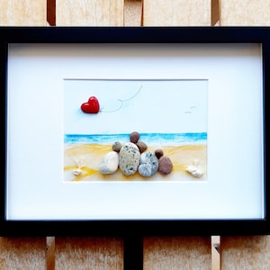 May include: A framed pebble art picture with a red heart balloon flying away from a group of pebbles arranged to look like people on a beach.