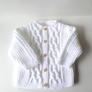 Puede incluir: Un cárdigan blanco de punto de cable con cuello redondo y mangas largas. El suéter tiene botones de madera en la parte delantera. El tejido de punto tiene un patrón texturizado y probablemente está hecho de hilo suave.